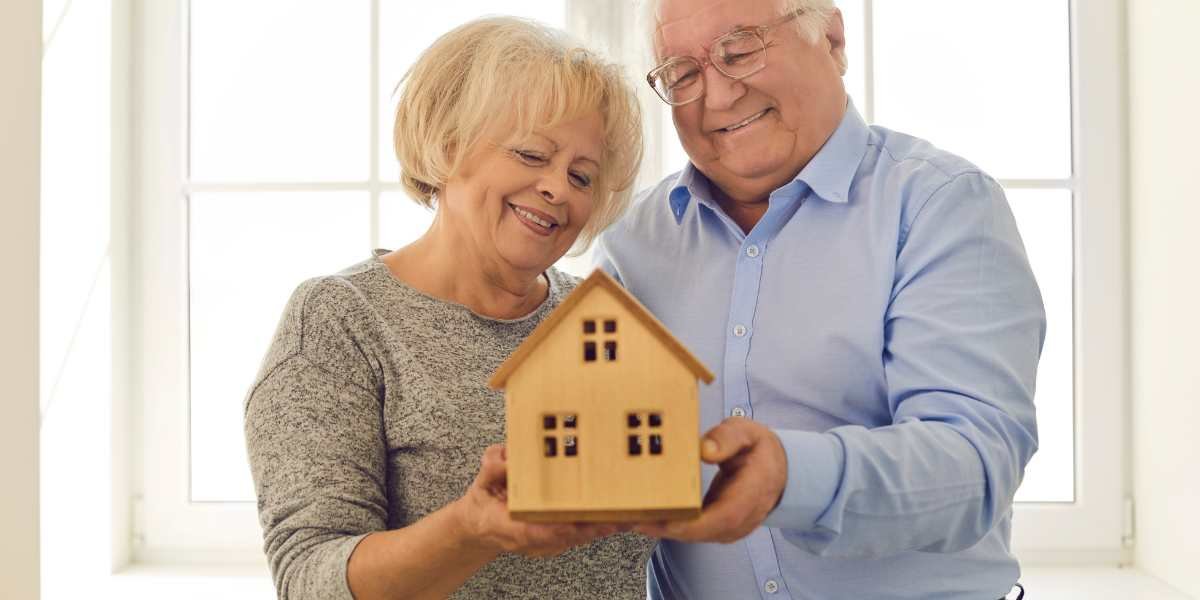 A happy senior couple smiling while holding a small wooden model house, representing over 50s life insurance, estate planning, and leaving an inheritance for loved ones.