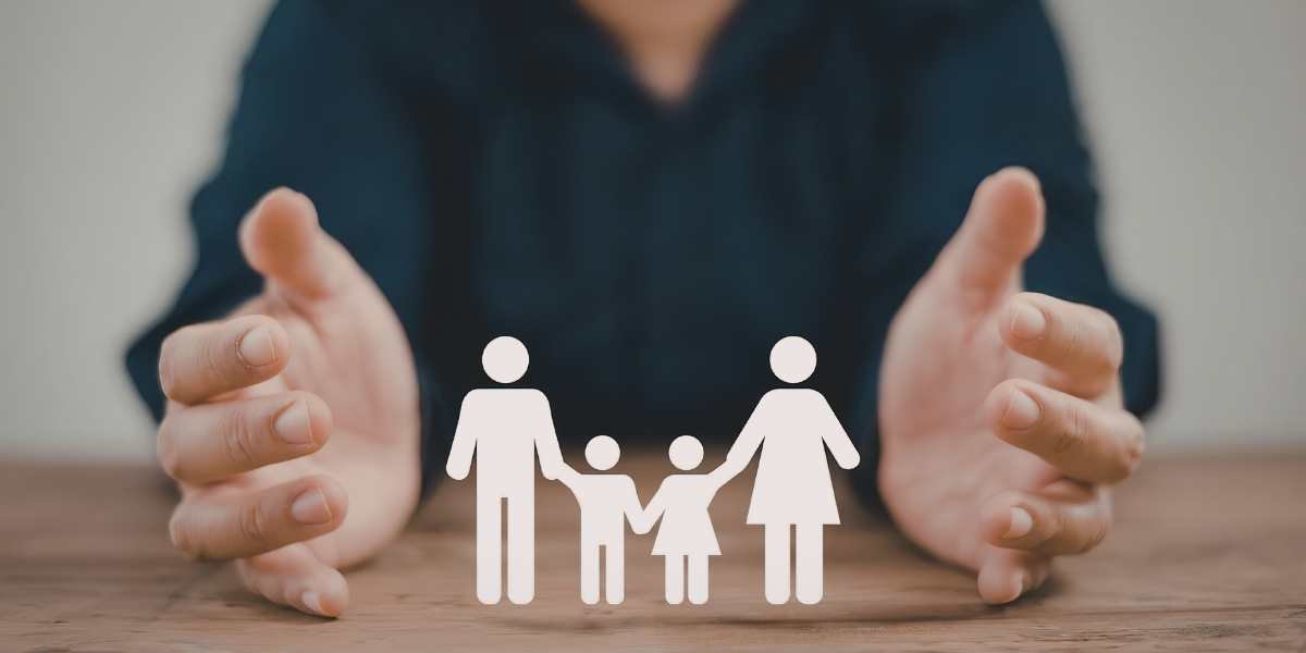 A person's hands framing and protecting a paper cutout of a family with two children on a wooden surface, representing family life insurance coverage in the UK.
