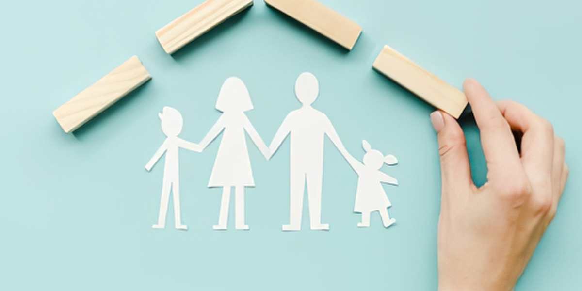 A hand arranging wooden blocks to build a protective roof over a paper cutout family on a light blue background, illustrating the building phase of life insurance coverage and qualifying waiting periods.
