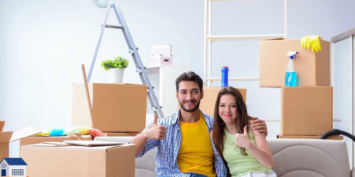 A smiling young couple sitting together on a sofa and giving a thumbs up, surrounded by stacked cardboard moving boxes, cleaning supplies, and a stepladder in their new home.