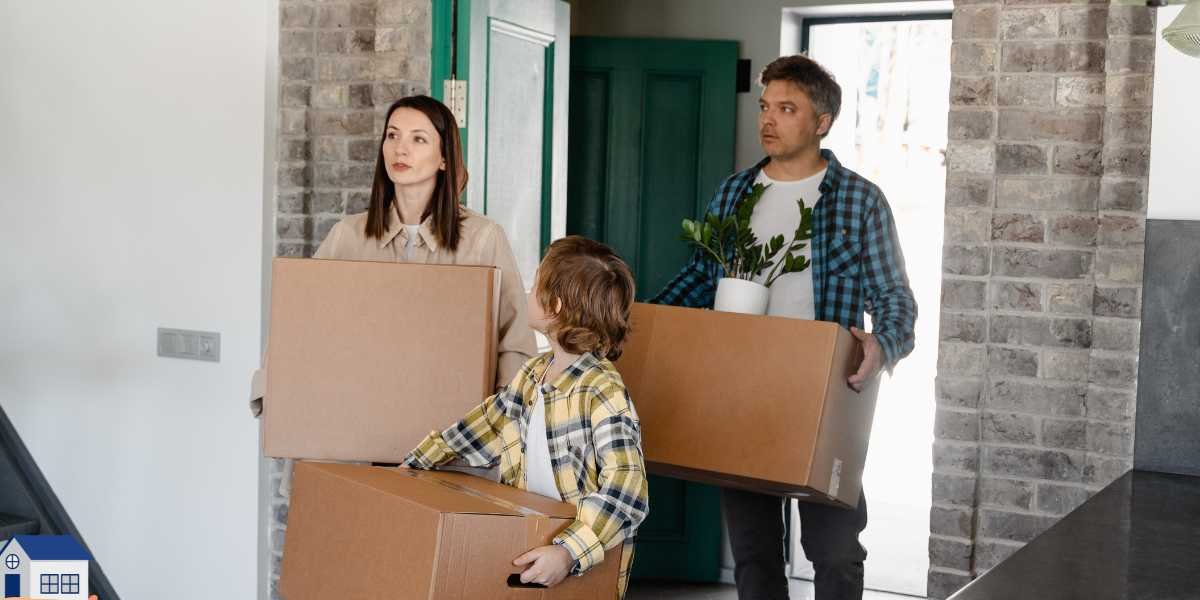 A family with a young boy carrying large cardboard boxes and a houseplant as they walk through the open doorway into their new home during the moving process.