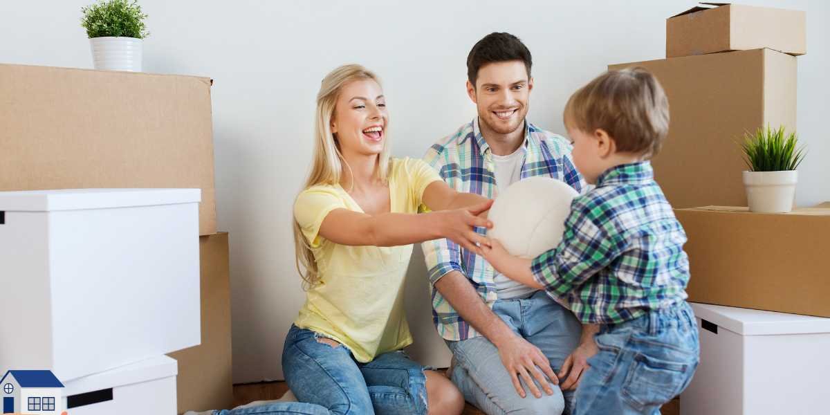 A happy young family sitting on the floor of their new home surrounded by moving boxes, with the mother laughing and reaching out to play with her young son.
