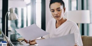 A self-employed professional businesswoman sitting at a desk with a laptop, carefully reviewing financial paperwork and tax documents for a mortgage application.