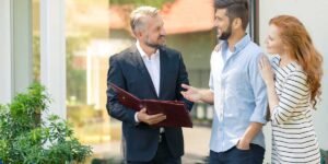 A smiling, professional real estate agent or financial broker in a dark blazer holding a maroon document folder outdoors, discussing options with a young couple—a bearded man in a light blue shirt and a woman with long red hair in a striped dress—who look pleased and engaged.