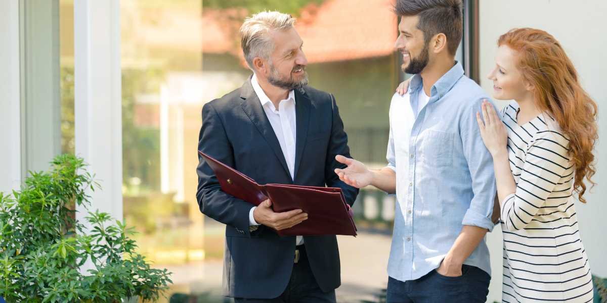 A smiling, professional real estate agent or financial broker in a dark blazer holding a maroon document folder outdoors, discussing options with a young couple—a bearded man in a light blue shirt and a woman with long red hair in a striped dress—who look pleased and engaged.