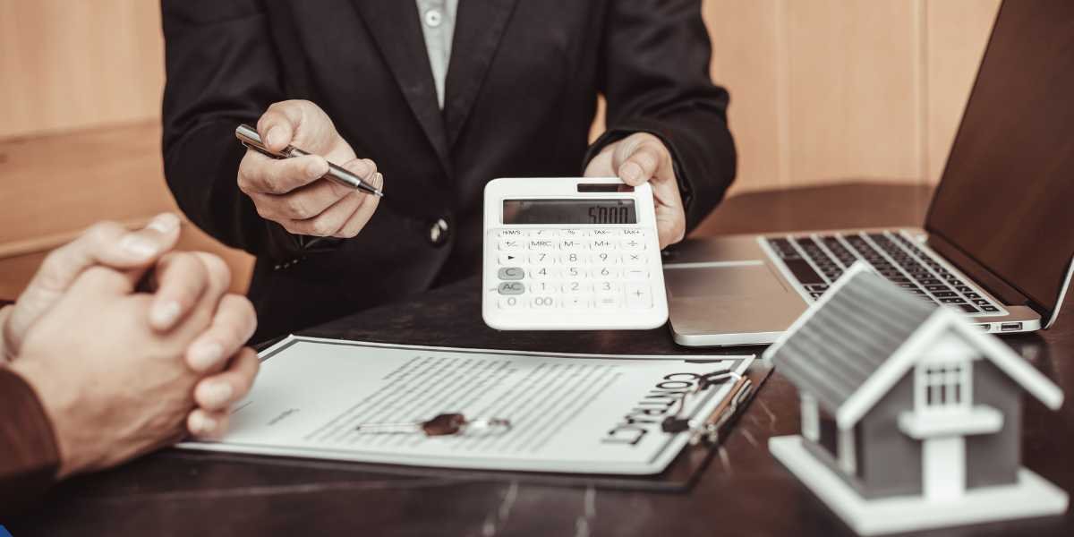Here is a tailored alternative text (alt text) for the image Nz Cover (1200 x 600 px) - 2026-06-23T165909.340.jpg, optimized for your UK property auction and bridging loan guide: Alt Text A financial specialist or mortgage broker in a business suit sitting at a desk, pointing a pen toward a property contract and holding out a white calculator showing a calculation. A small model house, a laptop, and a client's hands are visible in the foreground, representing a fast-tracked property auction finance consultation.