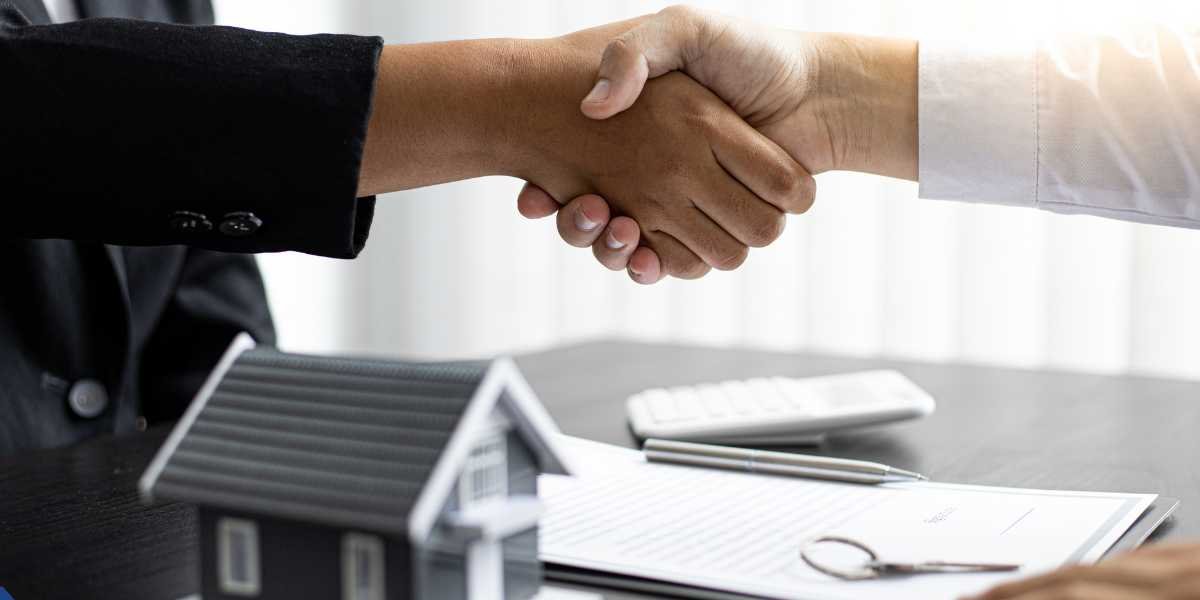 Close-up of a first-time landlord and a mortgage lender shaking hands over a desk with a small house model, calculator, and contract papers for property financing.