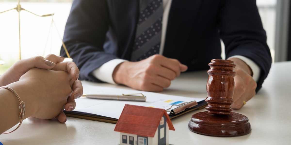 A close-up of a financial consultant or auctioneer in a suit holding a wooden gavel at a desk during a consultation. A small model house and a client's clasped hands sit in the foreground near a property contract, representing fast-paced UK property auction financing.