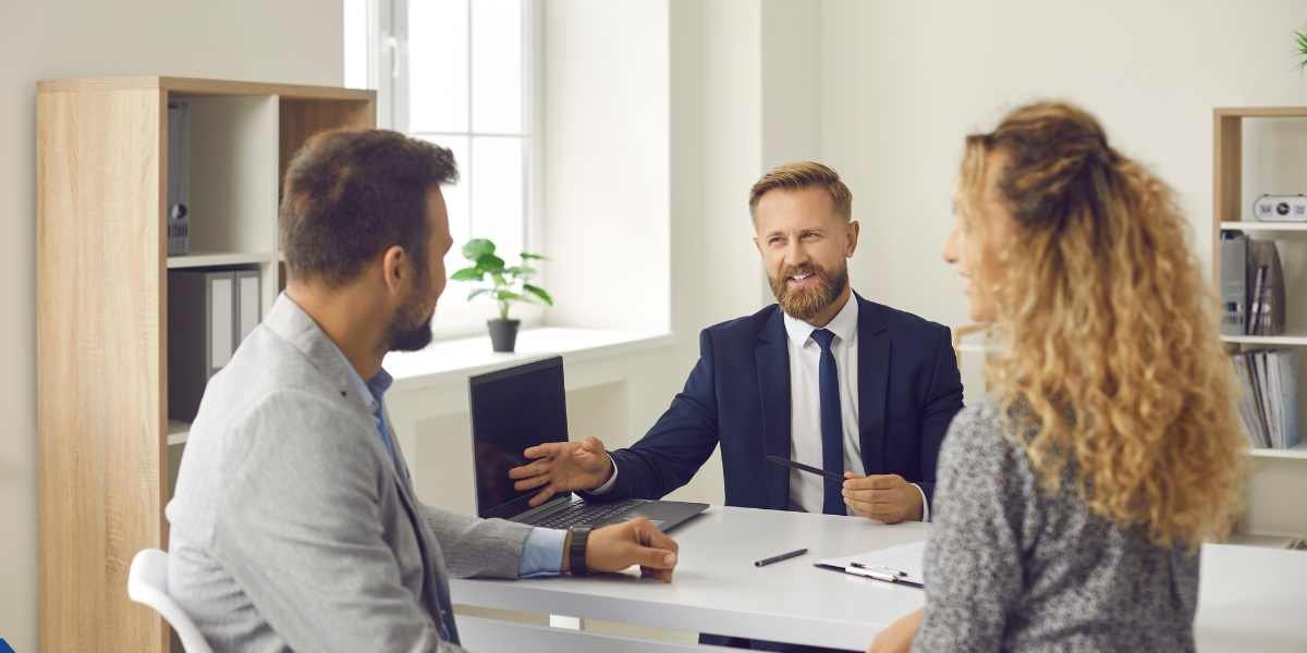 A bearded professional mortgage advisor in a suit explaining loan criteria and company profits on a laptop to a couple applying for a UK limited company director mortgage.