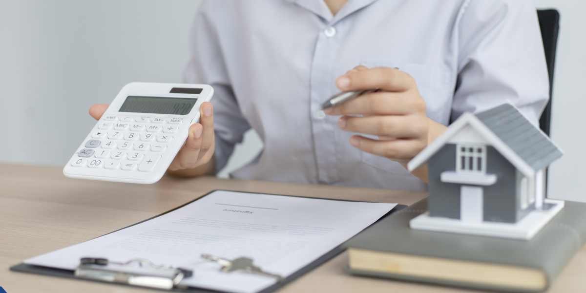 A close-up of a financial advisor or property specialist using a white calculator over a clipboard with a loan contract, with a small model house sitting on a book in the foreground, representing UK property auction finance calculations.
