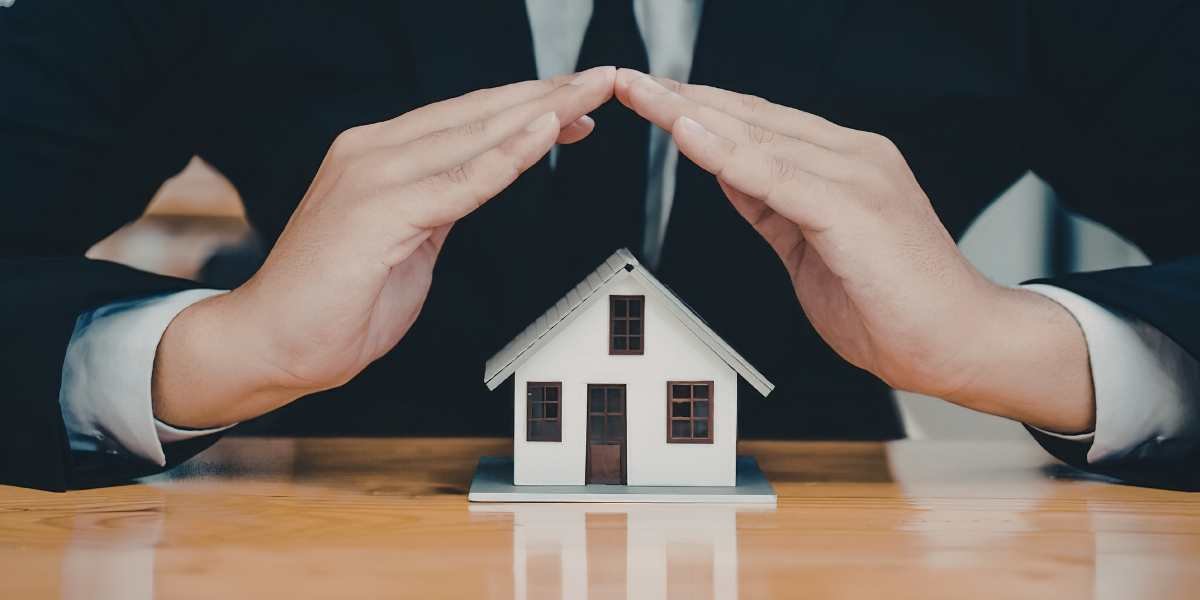 A person in a business suit cupping their hands over a miniature model house on a wooden desk, symbolizing consumer safeguards and property protection.