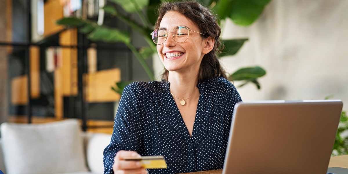 A smiling woman with glasses holds a credit card while looking at her laptop screen, representing an applicant reviewing her financial details online.