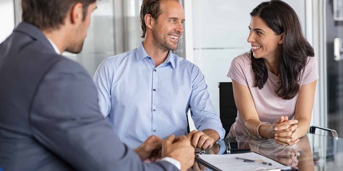 A smiling couple sitting at a glass office desk across from a financial advisor or mortgage broker in a grey suit jacket. They are engaged in a professional consultation with paperwork and a pen resting on the table in front of them, symbolizing a contractor mortgage advisory meeting.