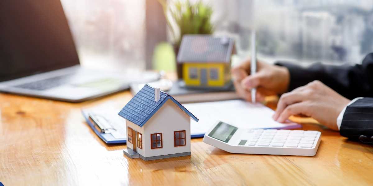 A small toy house model with a blue roof sits on a wooden desk next to a white calculator. In the background, a person's hands are visible signing financial documents next to an open laptop.