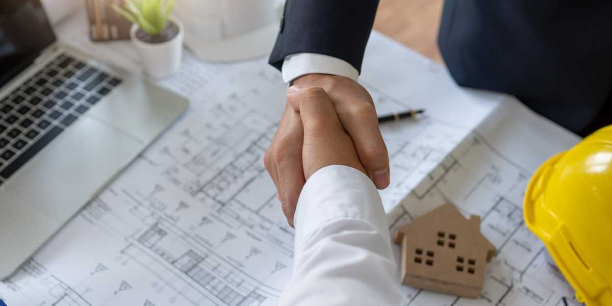 An overhead close-up of two business professionals shaking hands over architectural blueprints on a desk. A yellow construction hard hat and a small wooden toy house block are visible on the right.