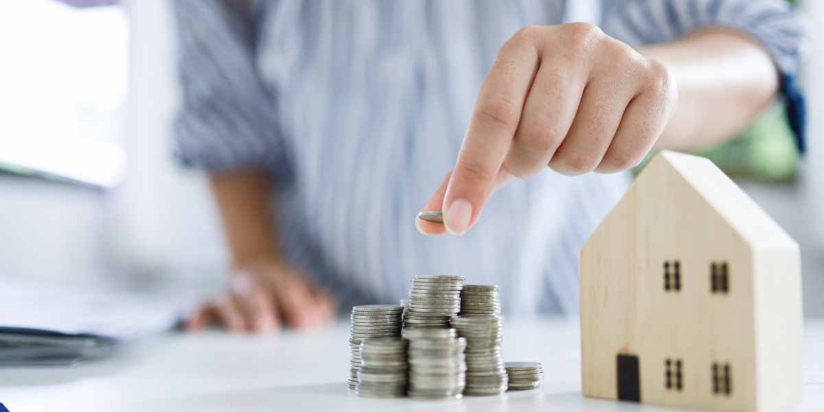Close-up of a person stacking coins next to a wooden toy house, representing how UK homeowners use home equity to manage and pay off multiple debts through mortgage consolidation.