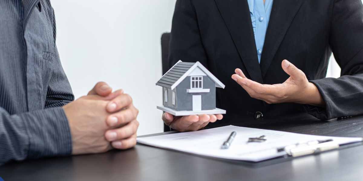 Close-up of a mortgage advisor holding a small house model across a desk from a client, demonstrating how debt consolidation mortgages work by using home equity to consolidate personal loans.
