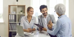 A young couple sits at a desk smiling and talking with an older female financial advisor in a bright office."