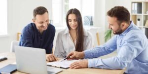 A young man and woman sit at a bright office desk, smiling as a mortgage advisor points to a contract with a pen, illustrating a Joint Borrower Sole Proprietor (JBSP) mortgage meeting where a family member helps a buyer purchase a home in the UK.