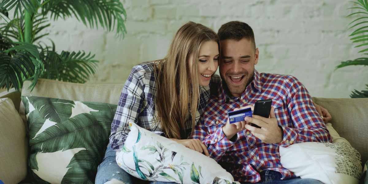 A young man and woman sit on a sofa, laughing while looking at a smartphone and holding a credit card, showing casual credit card usage.