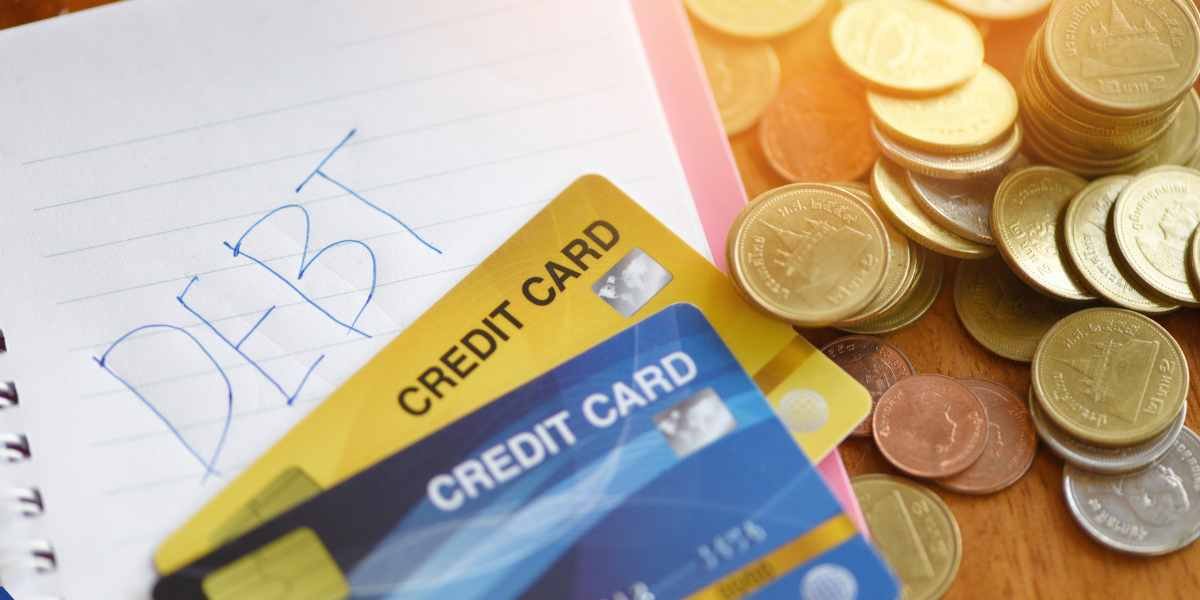 A notebook with the word 'DEBT' handwritten on it sits next to two credit cards and several stacks of gold coins, symbolizing credit card debt management when applying for a mortgage.