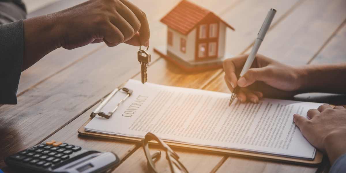 Close-up of a person signing a mortgage contract while another person hands over a set of house keys next to a toy home, representing a Joint Borrower Sole Proprietor arrangement where financial responsibility is shared but legal ownership belongs to one person.