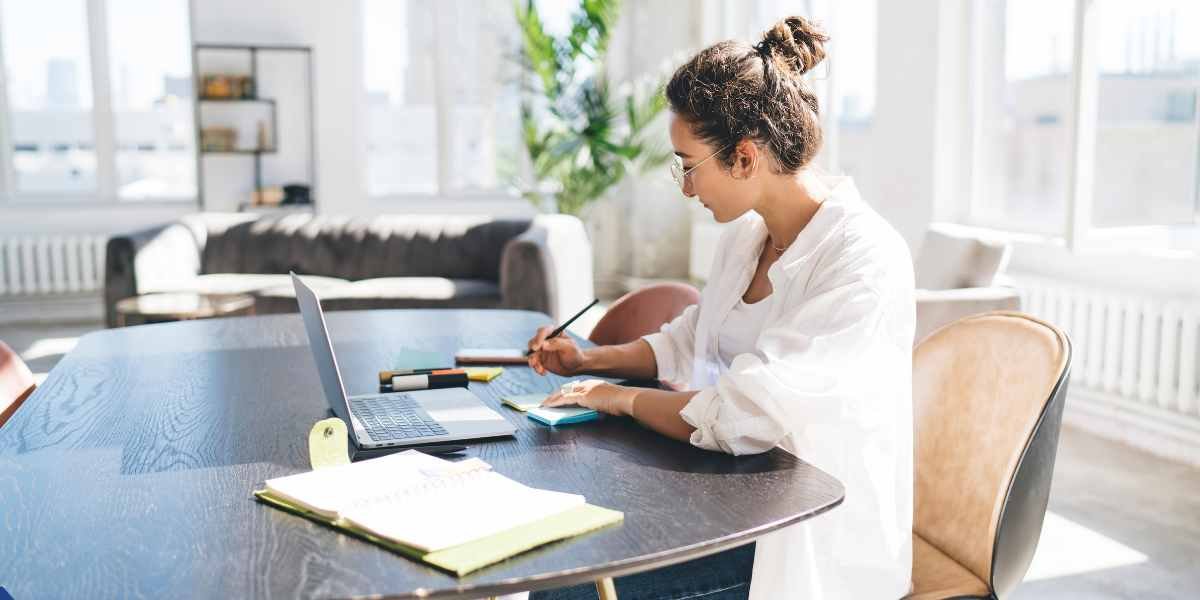 A young woman sitting at a table in a bright room, using a laptop and taking notes while researching first-time buyer mortgage schemes and property prices in Enfield.