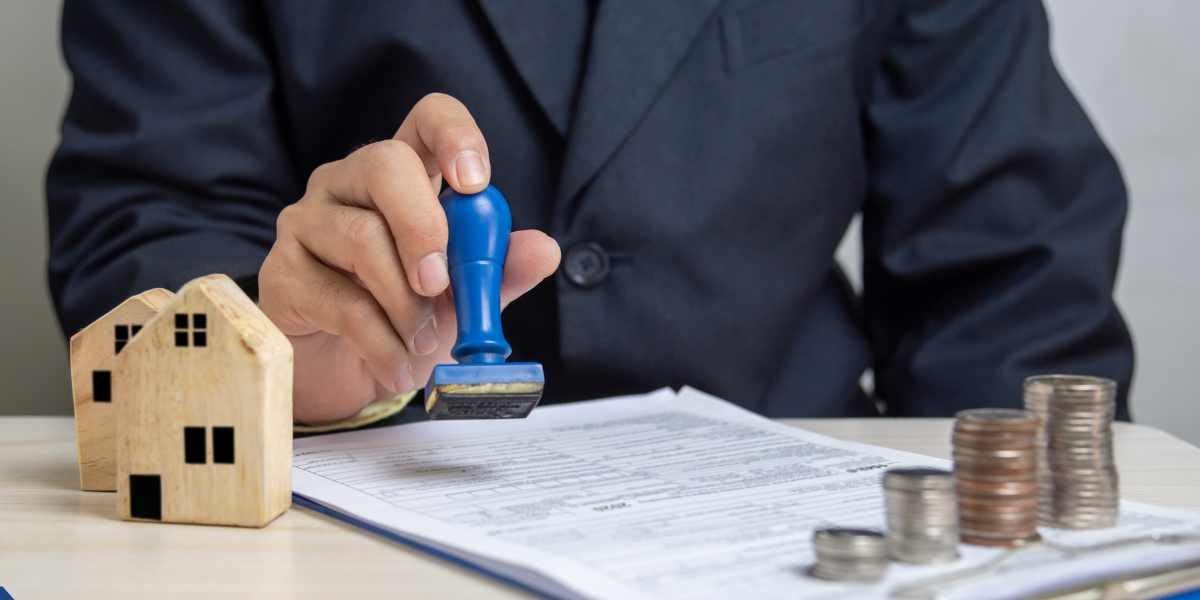 An advisor uses a blue stamp on a legal document next to miniature wooden houses and coin stacks, highlighting the formal approval process of a sole proprietor home loan.
