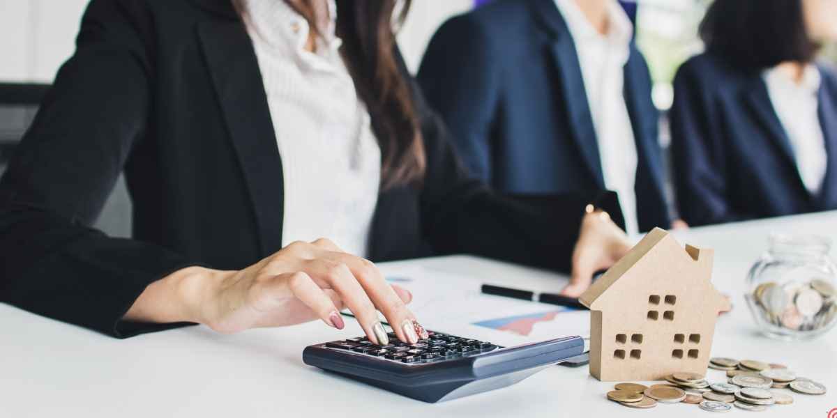 A business professional using a calculator next to a small wooden model house and stacked coins on a desk.